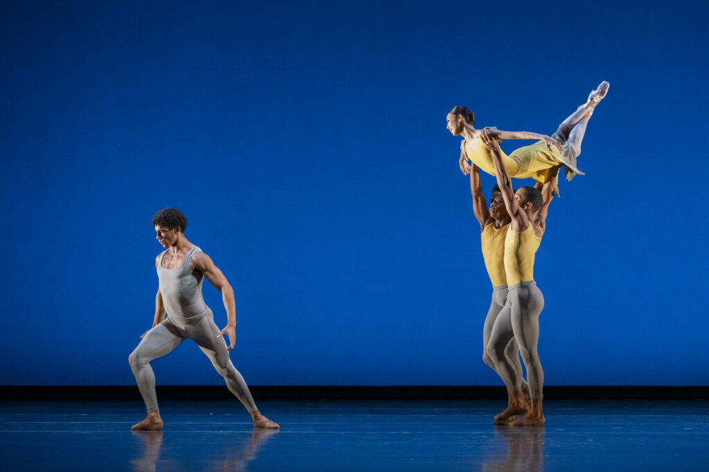 Onstage, one dancer lunges forward as a trio performs a group lift. The dancer on top arches up with her ankles crossed and arms gently extended to the sides. The two dancers lifting her support her by the elbows and knees as they walk forward. They all wear costumes with gray and yellow colors.
