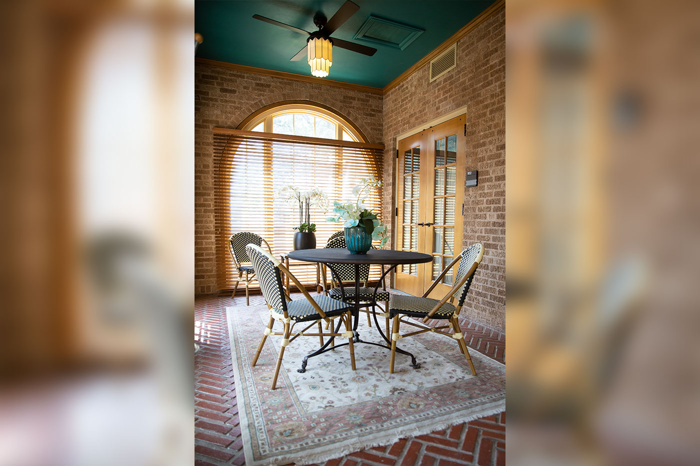 A cozy interior seating area features a round café table, four patterned chairs, potted flowers and a rug, set against brick walls with arched windows, wooden blinds and French doors.
