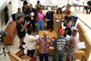 Group of diverse people standing in a circle in a living room, singing from songbooks with a camera and boom mic watching nearby.