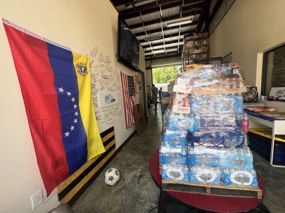 A pallet filled with bottled water and other drinks sit in a shipping facility garage with the Venezuela flag