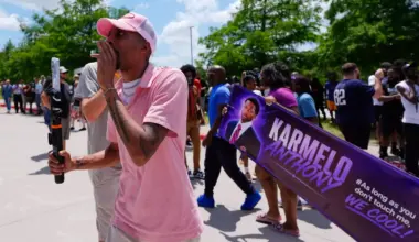 A man in a pink shirt holding a phone shouts into a crowd. Behind him is a banner that says "Karmelo Anthony"