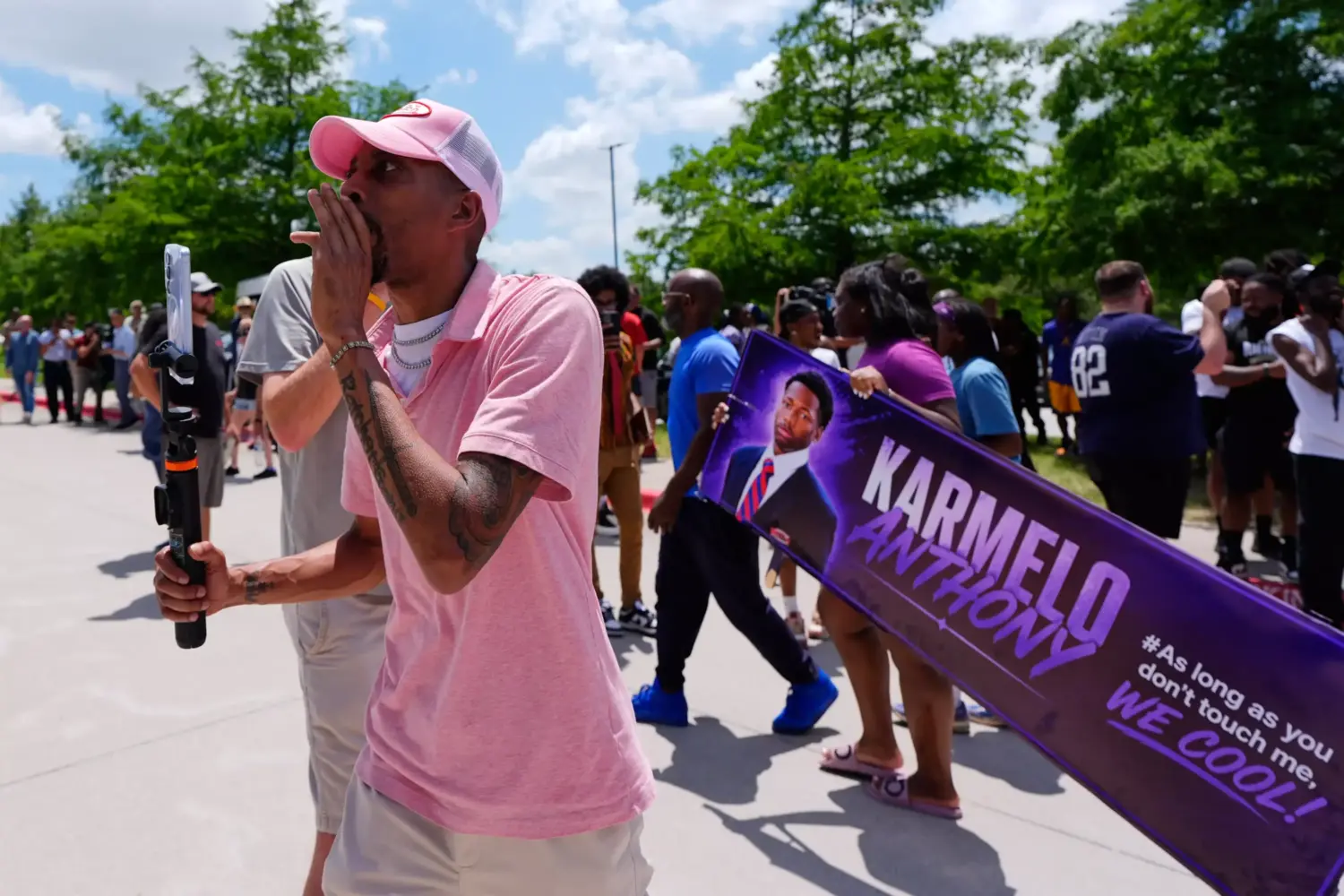 A man in a pink shirt holding a phone shouts into a crowd. Behind him is a banner that says "Karmelo Anthony"