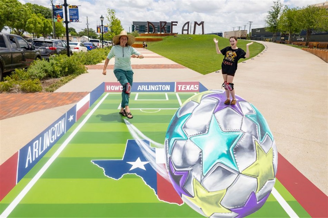 A person walks on a 3D artwork featuring a large, multicolored soccer ball on a Texas-themed field with 