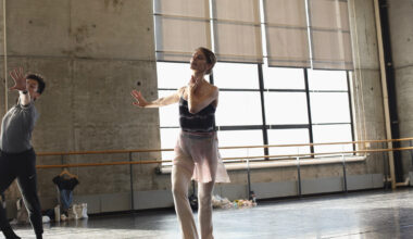In a warehouse-like ballet studio, Jacquelyn Long demonstrates movement, her hand on her neck, as another dancer follows.