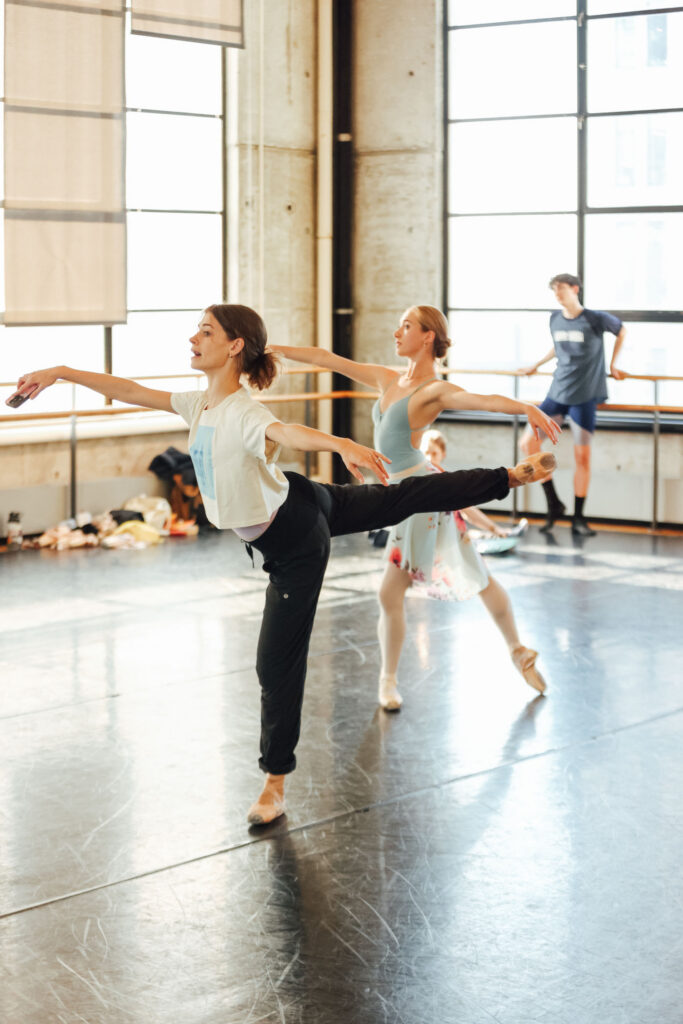 In a ballet studio, Jacquelyn Long demonstrates an arabesque with a slight fondu as a dancer stands in tendu derriere behind her. Another dancer leans on a barre watching.