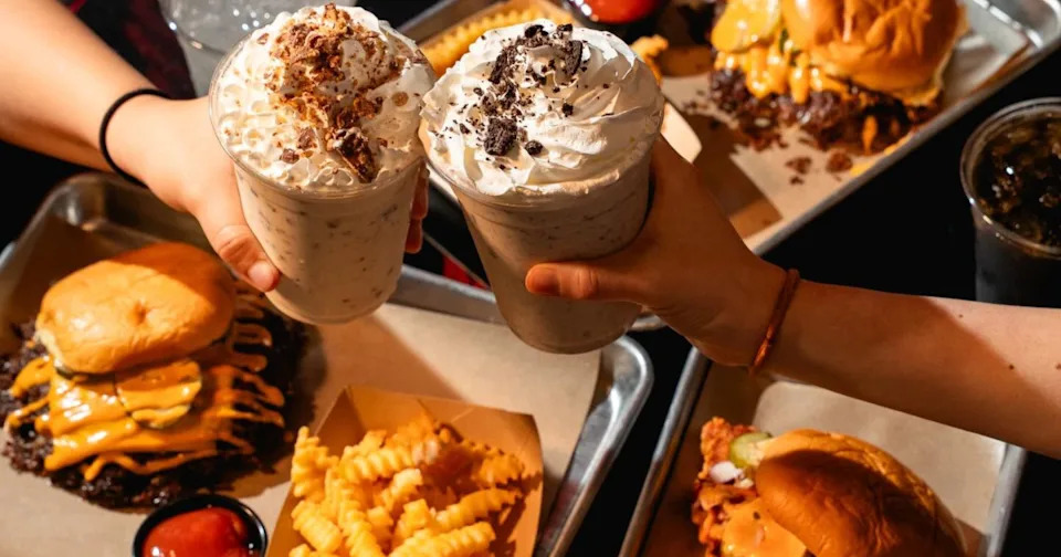 Two hands with shakes toast over trays of food from ZOJAH Burger in San Antonio
