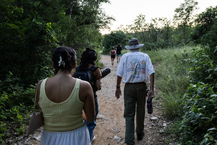 Three people walk along a trail outside at dusk. One person carries a microphone. 