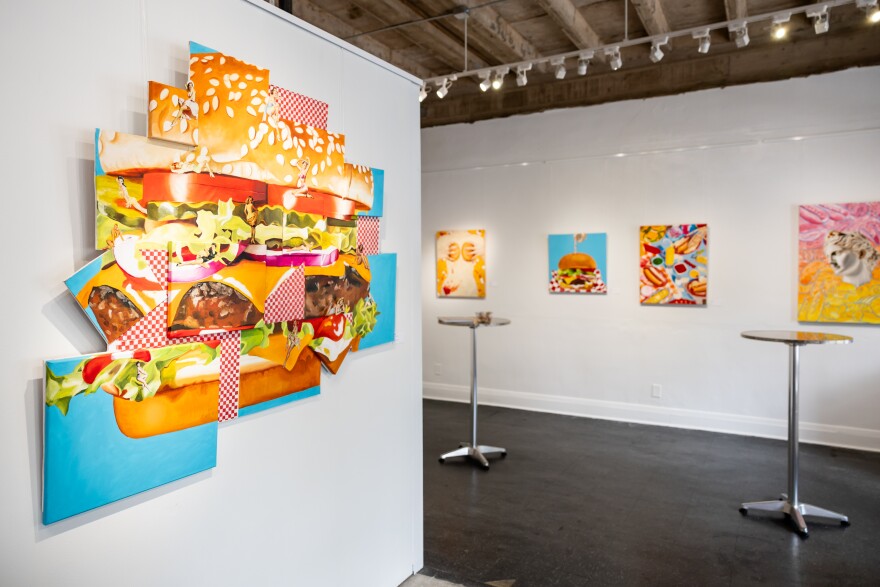 A painting that shoes a hamburger in panels hangs on a wall with tall tables and another gallery wall in the background.