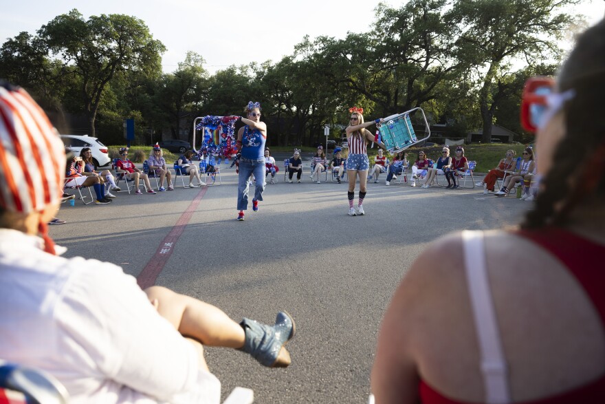 Women sit in the foreground and background watching two women in the center of a circle lifting lawn chairs over their heads. 