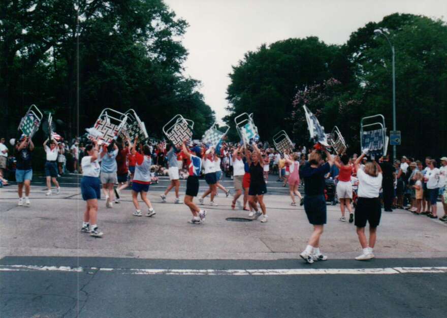 A grainy old photograph of a group of women marching in a parade with lawn chairs lofted overhead.