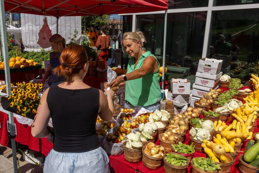 A woman at a booth at a farmer's market hands a bag of produce to another woman.