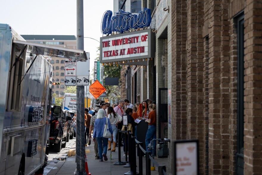 People line up on a city street outside a club. The club marquee is seen above the crowd.