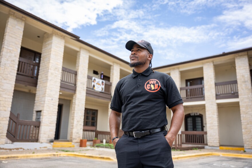 A man stands in front of a building posing for a photo with his hands in his pockets. He is looking up toward the left of the frame.