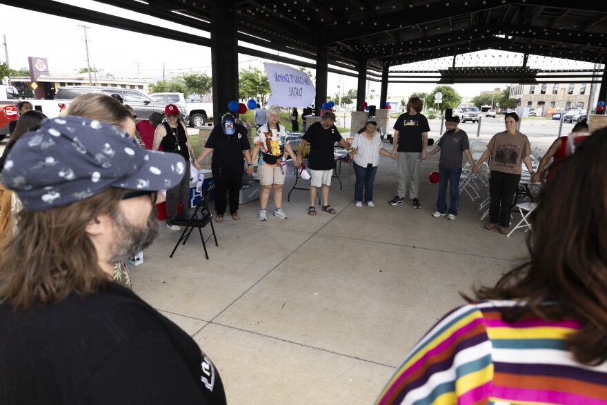 A group of people stand in a circle holding hands outside under an awning.