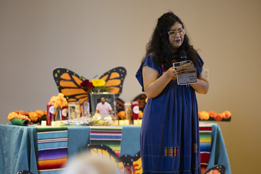 A woman with long hair and a blue dress speaks to an audience in front of a memorial shrine.