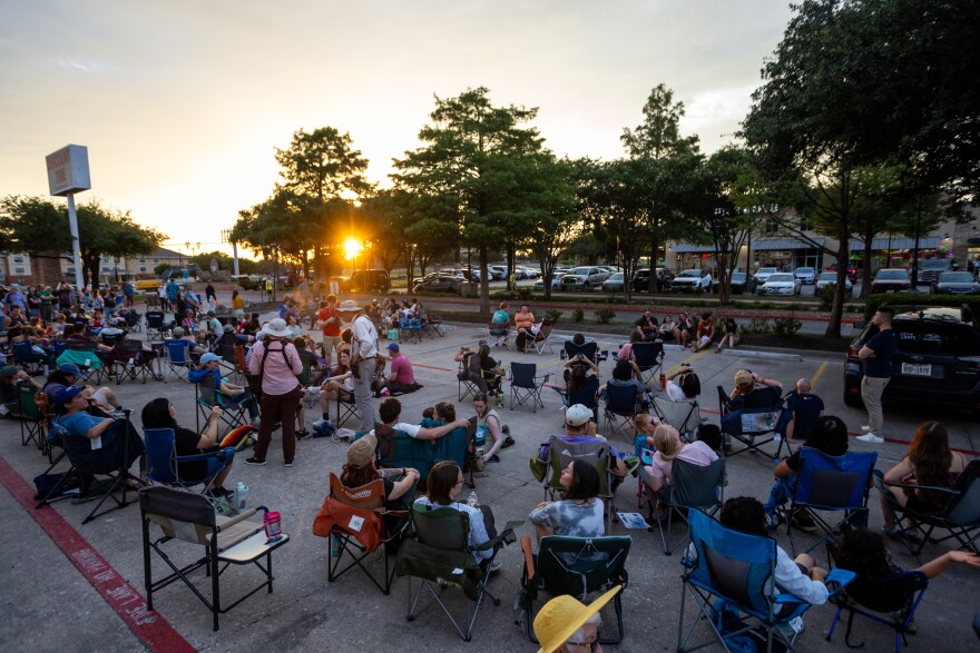 A large crowd sits in foldable chairs in a parking lot as the sun sets.