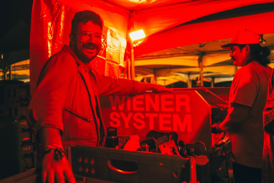 A man smiles for the camera as he stands next to a cart that says "wiener system."