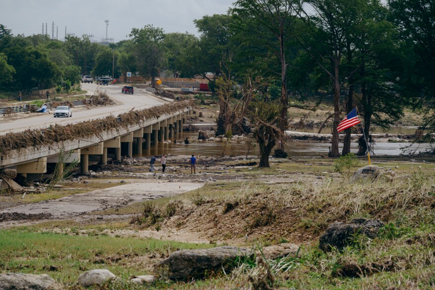 A bridge is seen to the left near a river and a park with obvious signs of flooding including knocked over vegetation. A U.S. flag flies on a pole to the right.
