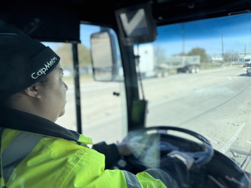 A bus driver with a black "CapMetro" knitted hat and neon safety vest operates the steering wheel of a bus. You can see out the front windshield at some of the traffic head.