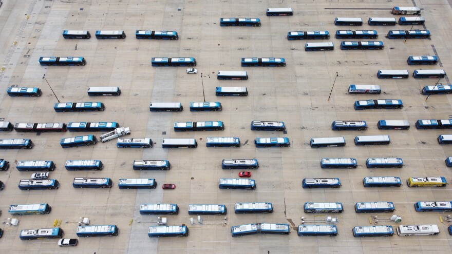An aerial view of CapMetro buses, both electric and diesel, parked at the North Ops Center. The buses are arranged in rows across a large, concrete parking lot, with a few small vehicles and maintenance equipment visible among them.