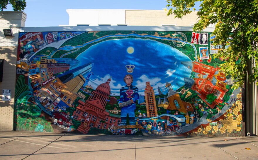 A mural that is painted with a fish eye perspective of a city is seen on a wall with a walkway in front and trees at the top sides.