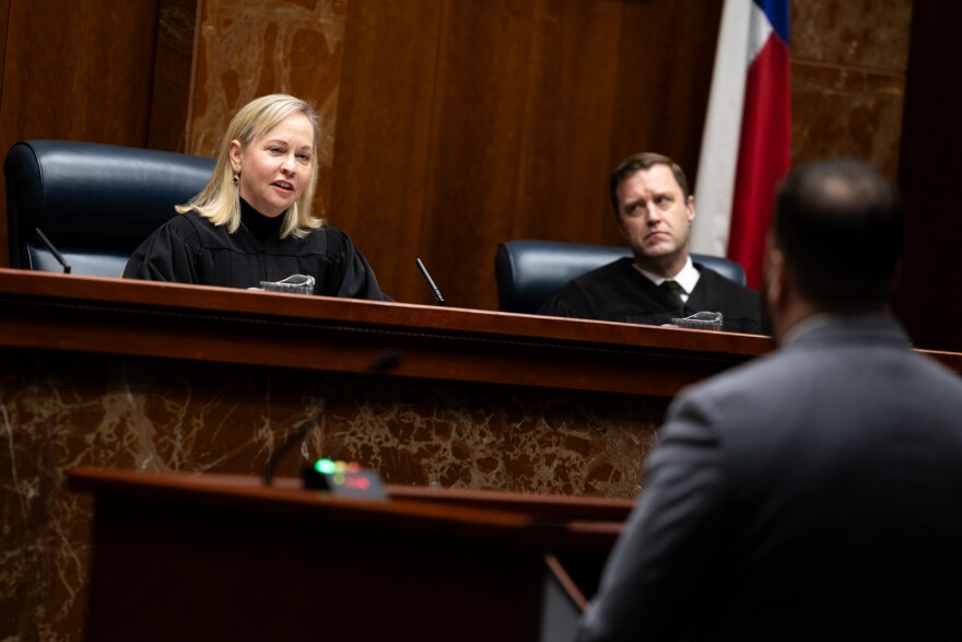 A judge sits at her bench and asks a question of a lawyer while another judge looks over at her. 