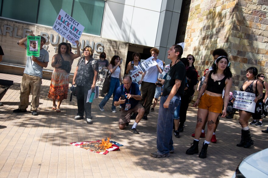 Protesters gather outside of an office building near a U.S. flag that is burning on the ground.