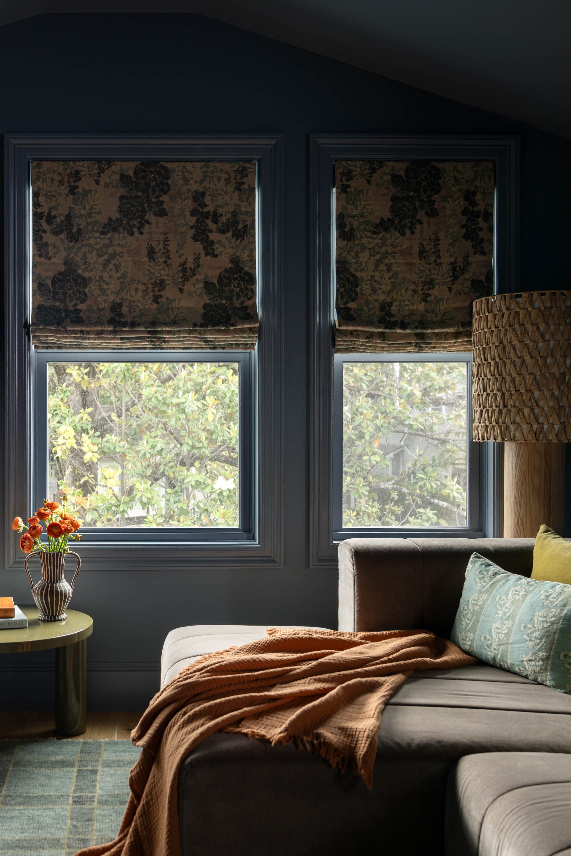 Cozy living room with a sectional sofa, an armchair, decorative pillows, and floral window shades. A vase with flowers on a table.