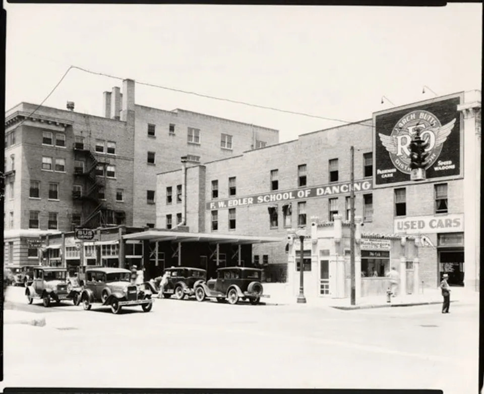 Looking northwest in 1931 from the intersection of First Street and Lawrence (now known as Broadway) in Wichita, Kansas. Businesses visible include: White Castle Hamburgers, Union Bus Depot, Tostwich Cafe, Butts New & Used Cars, Edler School of Dancing. The automobile in the left foreground appears to have an "Air Capital" emblem mounted on the grille.