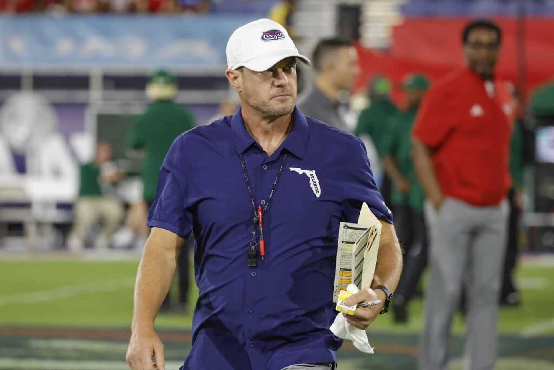 Tom Herman holds a play sheet and walks on the field wearing a Florida Atlantic shirt and hat