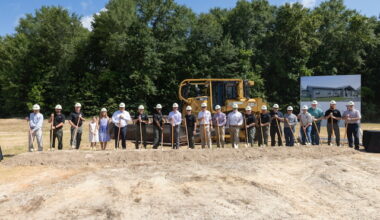 Twenty-one individuals wearing hard hats stand in front of a dozer while holding shovels to break ground on a new facility.