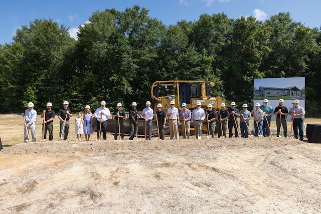 Twenty-one individuals wearing hard hats stand in front of a dozer while holding shovels to break ground on a new facility.