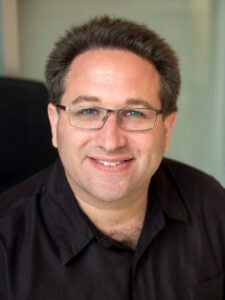 headshot of a man in a black shirt wearing glasses