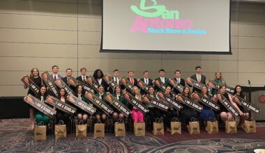A large group of students hold banners reading "I am a scholar." Behind them a screen displays the logo of the San Antonio Stock Show and Rodeo.