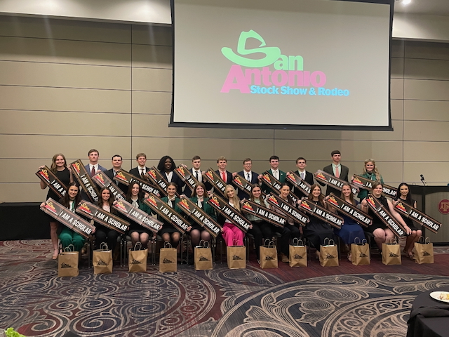 A large group of students hold banners reading "I am a scholar." Behind them a screen displays the logo of the San Antonio Stock Show and Rodeo.