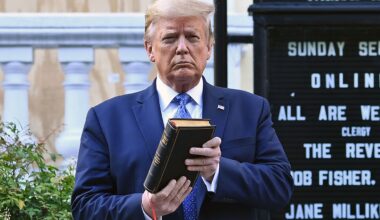 Trump holds up a closed book in front of a church sign outdoors. Shrubs and part of a building are visible in the background.