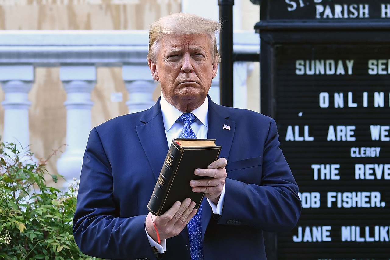 Trump holds up a closed book in front of a church sign outdoors. Shrubs and part of a building are visible in the background.