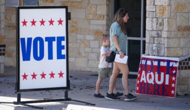 A woman and a child walk past large VOTE signs, one in English and one in Spanish, outside a stone building serving as a polling place.