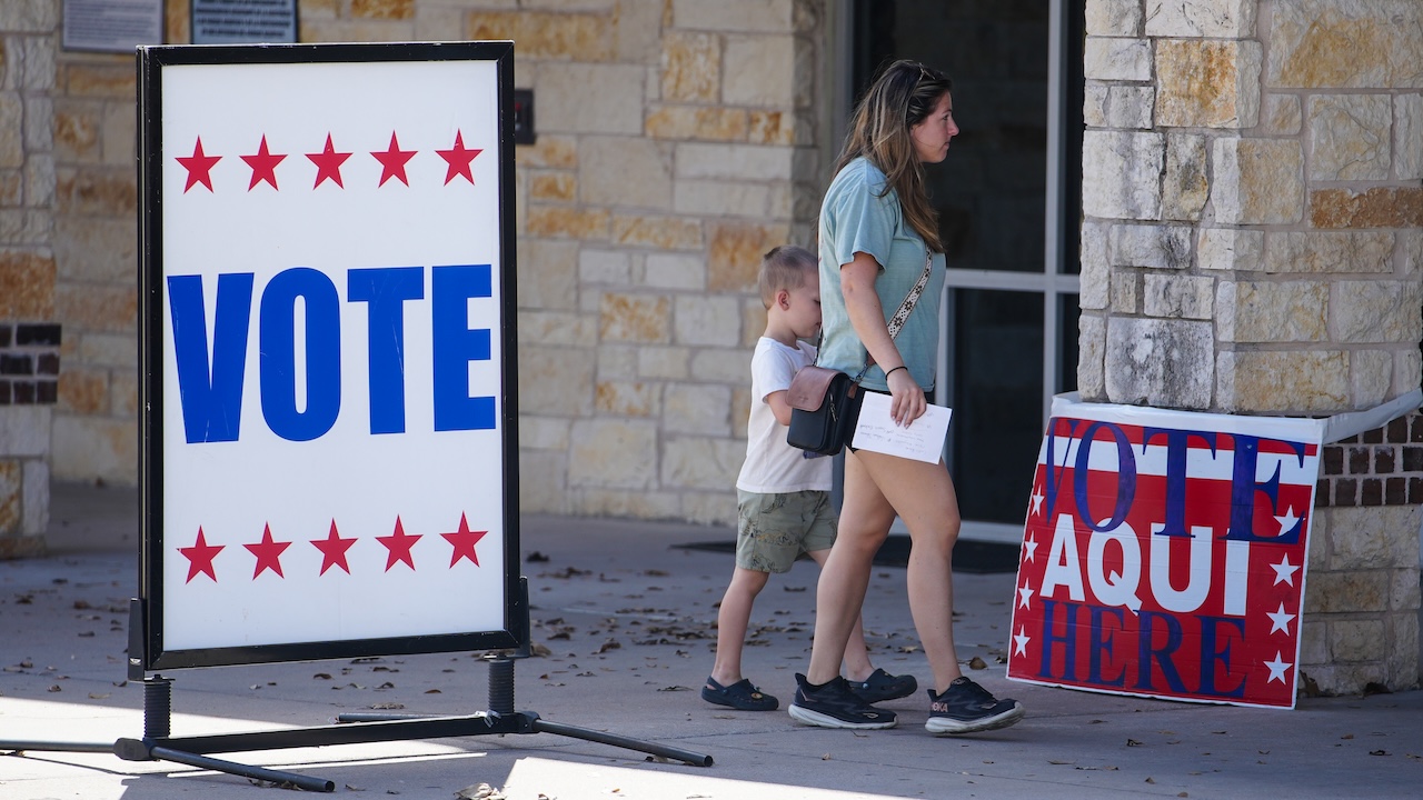 A woman and a child walk past large VOTE signs, one in English and one in Spanish, outside a stone building serving as a polling place.