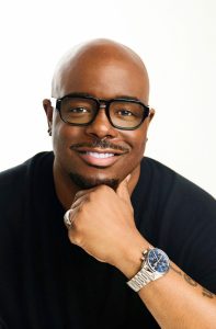 A headshot of John Gibson. He is wearing a black shirt and glasses. He is smiling in front of a white background.