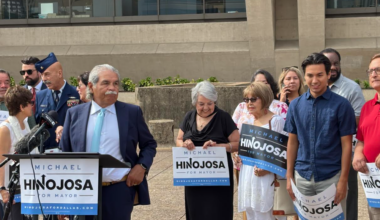 Michael Hinojosa stands at a podium outside Dallas City Hall in a suit and blue tie. Family, veterans and supporters stand behind.