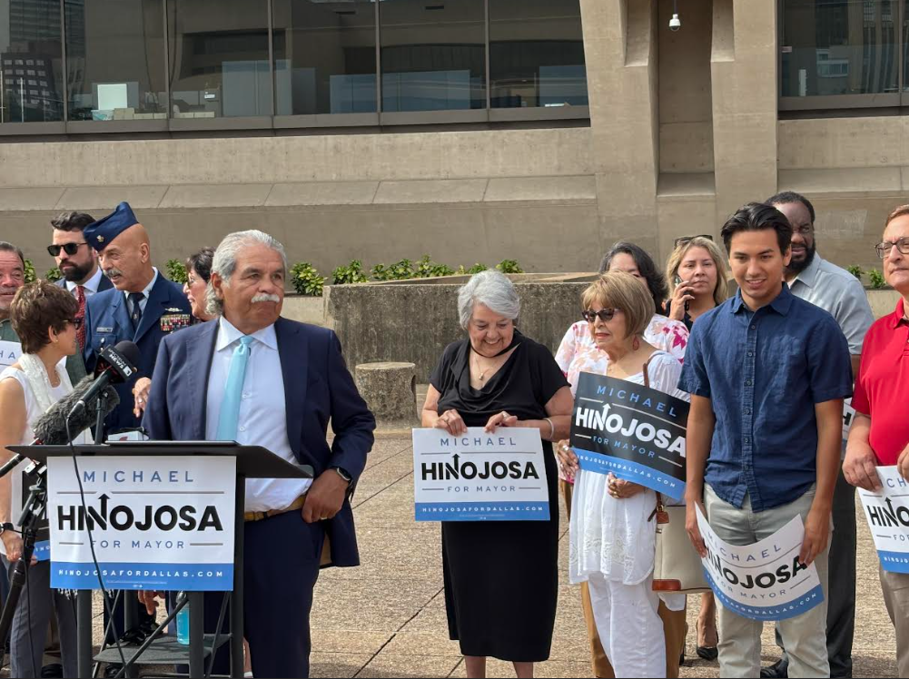 Michael Hinojosa stands at a podium outside Dallas City Hall in a suit and blue tie. Family, veterans and supporters stand behind.