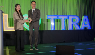 a woman wearing a brown jacket accepts an award from a man in a suit and the letters TTRA can been seen lit up in blue on the stage