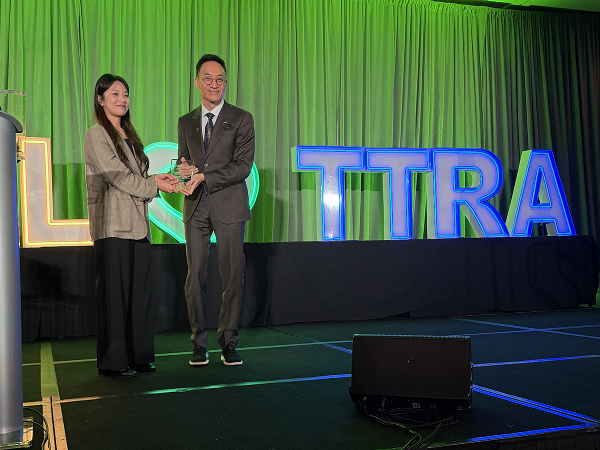 a woman wearing a brown jacket accepts an award from a man in a suit and the letters TTRA can been seen lit up in blue on the stage