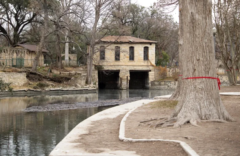 The historic pump house and Lambert Beach at Brackenridge Park are flanked by retaining walls that are crumbling due to encroaching tree roots and other factors. Some trees mislabeled by activists to be removed sit across the river.