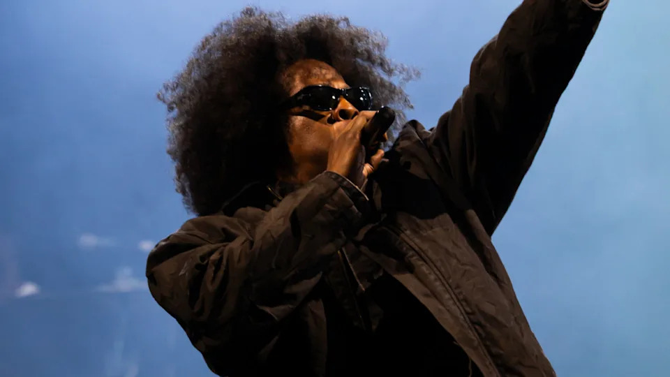 Jay-Z, with an afro and sunglasses, raps passionately on stage, wearing a dark jacket against a blue-lit background.