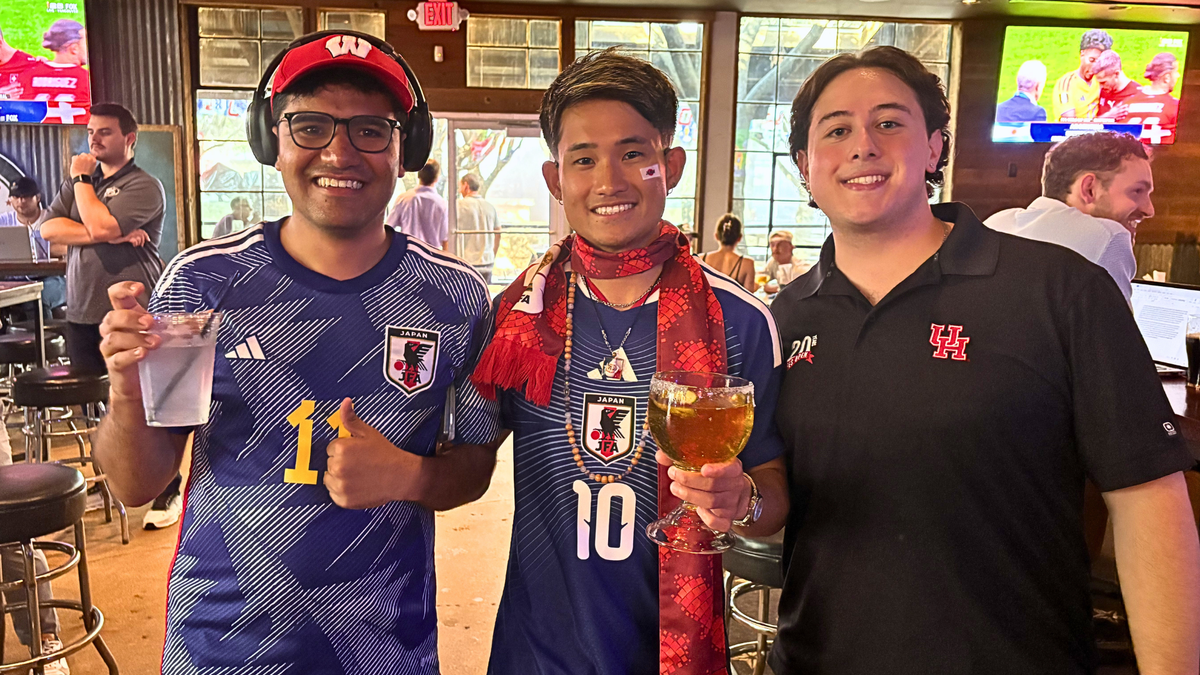Japanese social media influencer poses with soccer fans to his left and right at a Dallas bar, smiling while holding drinks, posing to the camera.
