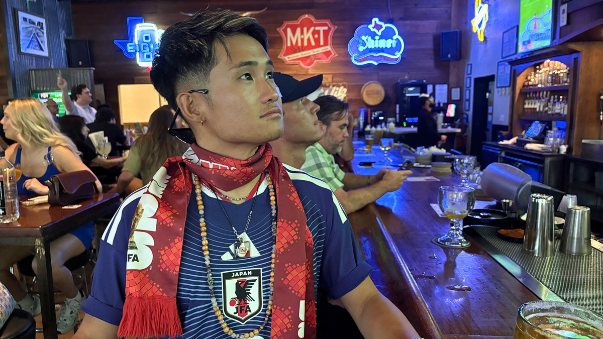 Asian man looks away at tv across from him while sitting at bar in Texas with neon signs in the background