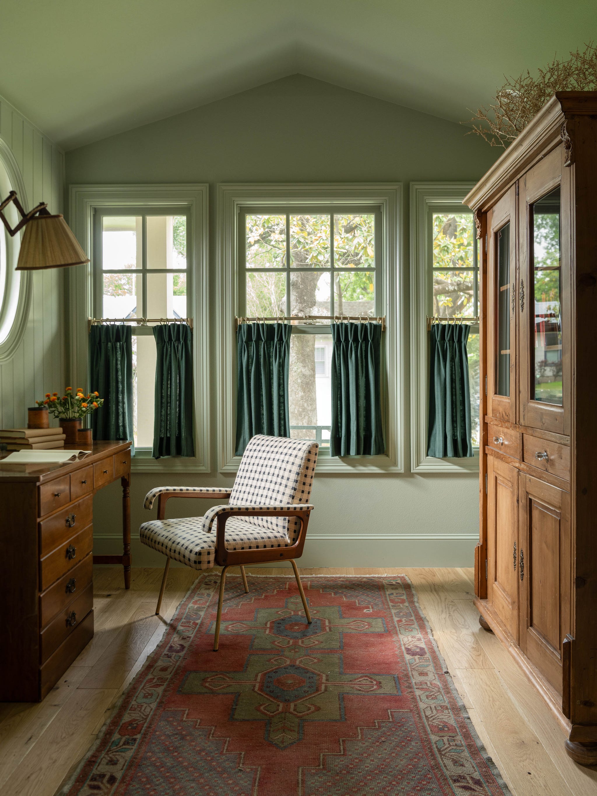 A cozy study corner featuring a wooden desk, a patterned chair, green curtains, and warm wooden accents.