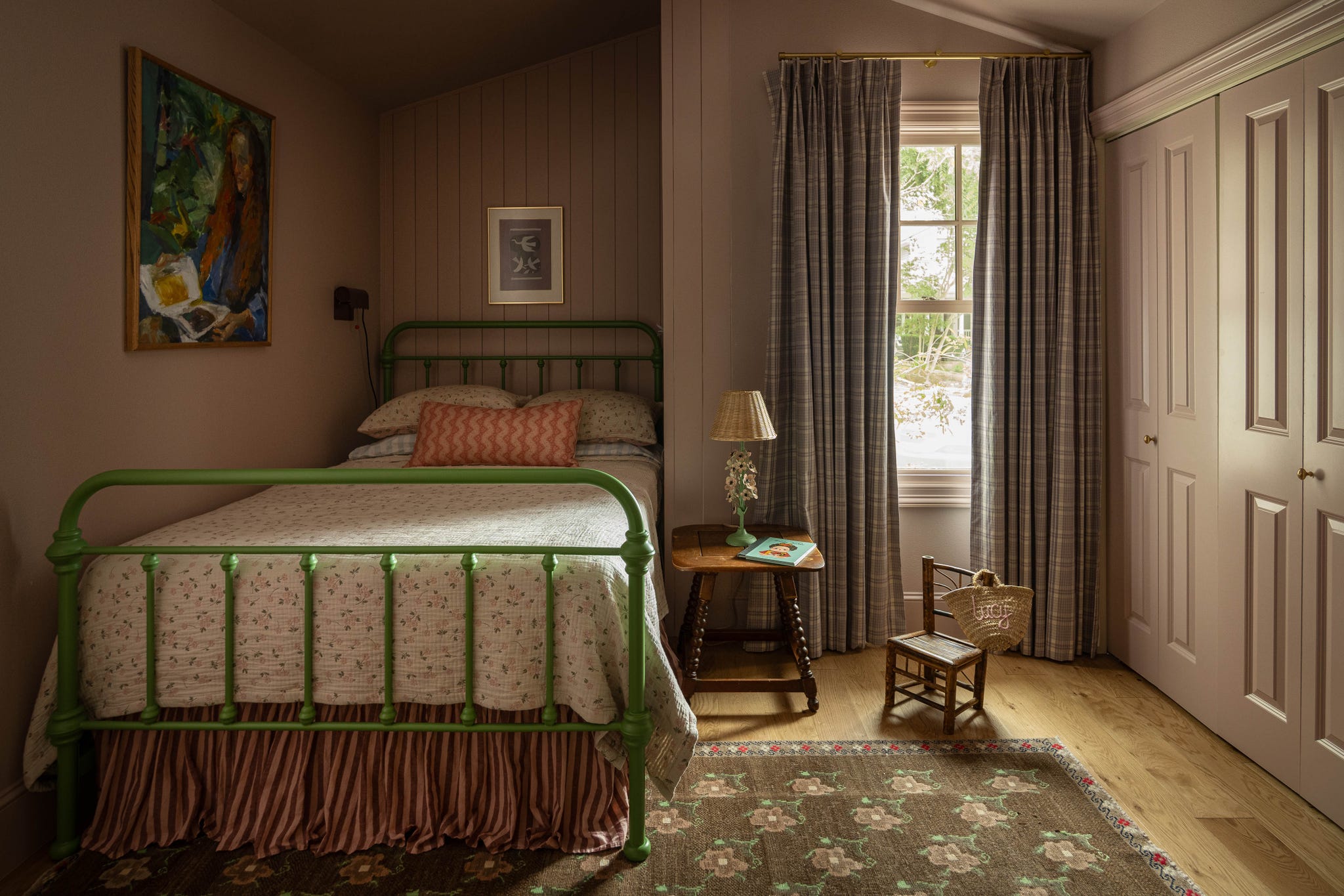 Cozy bedroom featuring a green metal bed, patterned quilt, a small wooden table, and large window with curtains.